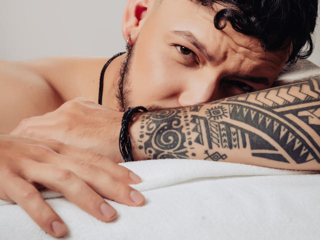 JaxRagnar with curly black hair, tattooed arm, and black necklace, resting his face on a white towel, showing a focused, slightly annoyed expression. His hand is on the towel.
