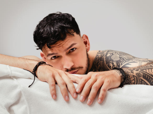 JaxRagnar, shirtless, with dark curly hair and tattoos, leans on a white surface, hands in front, brown eyes looking at camera, black bracelet on right wrist.