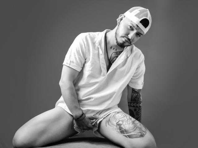 JaxRagnar, in a white shirt, cap, and no pants, sits with legs spread, holding his cock, tattoo on thigh, looking at camera with a slight smile, grayscale photo.