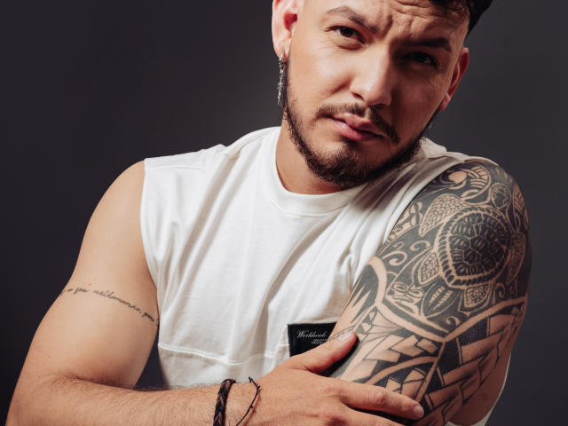JaxRagnar with a tattooed shoulder, white sleeveless shirt, and a beard, looking sideways with a slight smirk. His right hand touches his chest. Dark background.