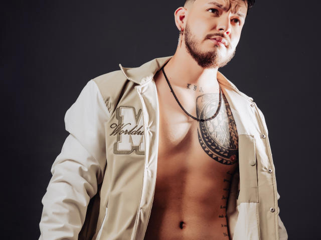 JaxRagnar, shirtless, with a toned, muscular chest, a large tattoo over the right pectoral, and a necklace, wearing an unbuttoned beige jacket, dark background, looking slightly to the side.