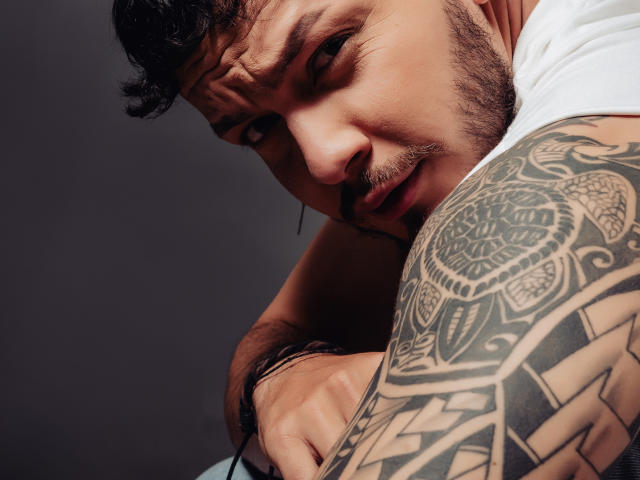 JaxRagnar with a tattooed arm, looking down with a slight smirk, short curly black hair, and a trimmed beard, wearing a white shirt, showing a muscular upper body.