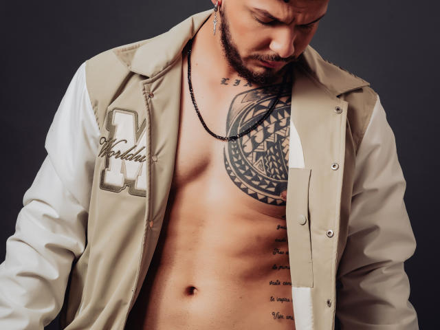 JaxRagnar's muscular chest, tattooed with a large circular design, is exposed under an unbuttoned beige and white varsity jacket. He has a beard and wears a black necklace.