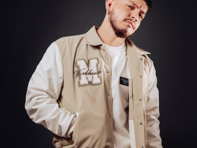 JaxRagnar with a beige varsity jacket, white shirt, hands in pockets, looking to the side, dark background, short beard, slightly tilted head, brown hair, casual style.