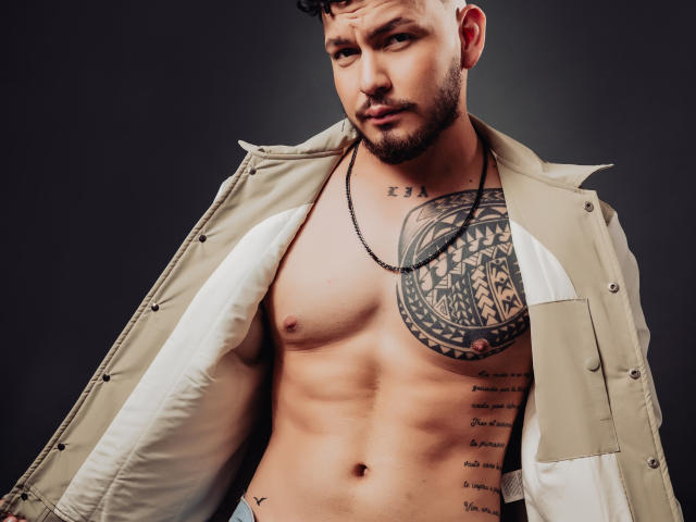 JaxRagnar has a toned, shirtless body with a large tattoo on his left chest and a chain necklace. He's wearing an open, beige jacket with a white shirt.