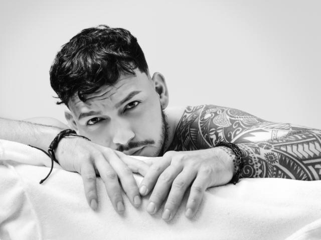 JaxRagnar, shirtless with tattoos on his right arm, leans on a white surface, eyes looking up, short dark hair, bracelet on left wrist, black earbuds, slightly wavy eyebrows.