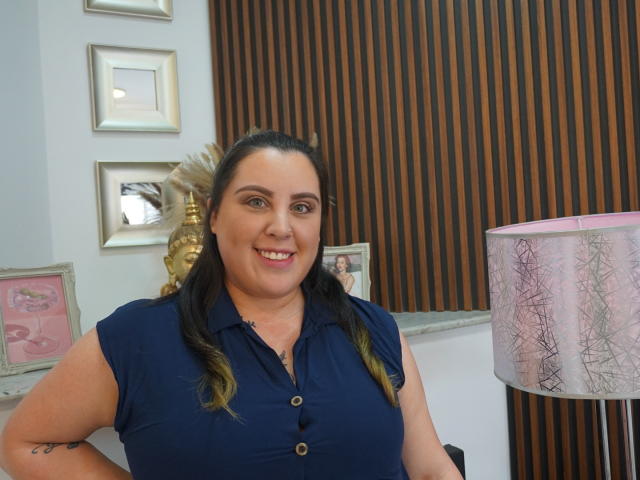 AgathaSantori met borsten in een marineblauwe blouse, lachend, bruin haar in staarten, roze lippenstift, tatoeage op de rechterarm, staand naast een roze lamp met een draadontwerp..
