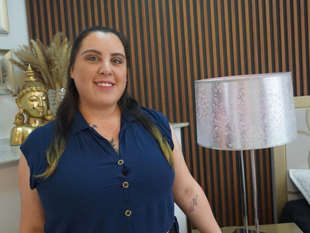 Agatha Santori, con los senos visibles debajo de una camisa azul marino, sonriendo, cabello castaño con puntas verdes, un tatuaje pequeño en el brazo derecho, de pie junto a una lámpara de color rosa con estampado..