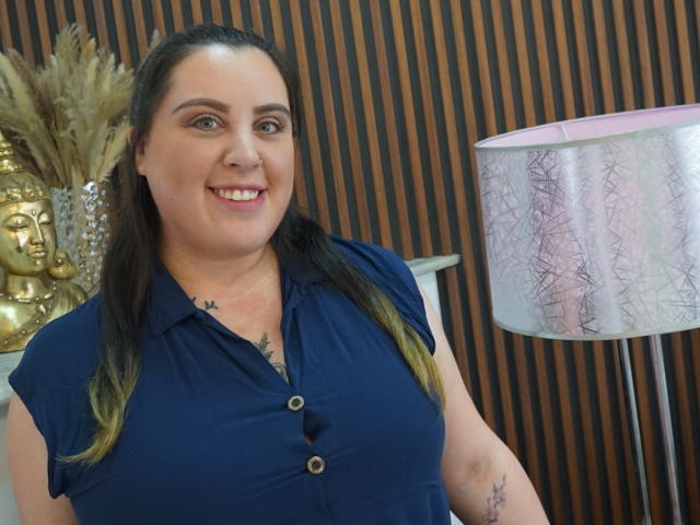 Agatha Santori mit mittelgroßer Brust, hellblaues Hemd mit Knopfleiste, Tattoos an Brust und Arm, braunes Haar, lächelnd, stehend neben einer pink-weißen Lampe mit Muster, Holz- und Golddekor..