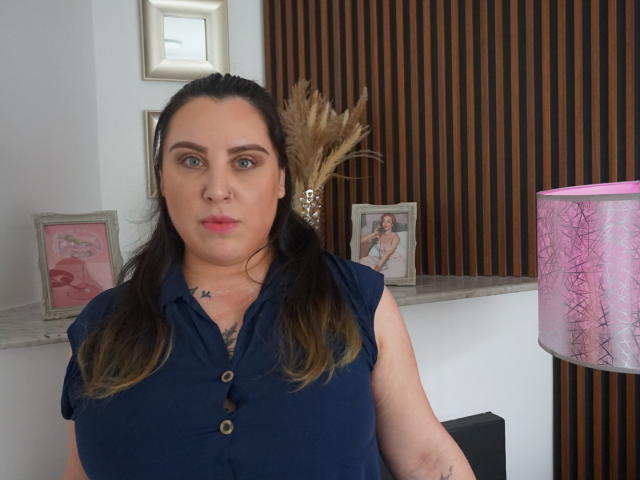 Agatha Santori, con cabello castaño, camisa azul abotonada, con los senos visibles, tatuajes en el pecho, mirando a la cámara, una lámpara rosa con un patrón blanco, una pared de madera, cuadros enmarcados detrás..