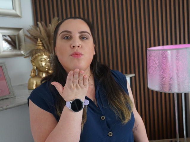 Agatha Santori, con cabello castaño, camisa azul, reloj de pulsera negro, mano cerca del rostro, con una expresión ligeramente molesta, en una habitación con una lámpara rosa y una estatua dorada de Buda..