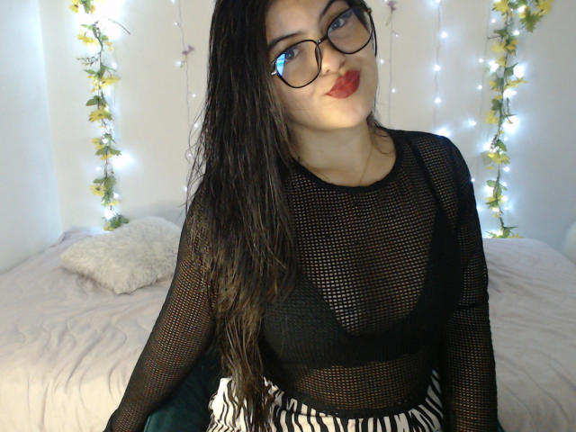 AlissonS with wet black hair, glasses, and red lipstick, wearing a sheer black mesh top showing her tits, and striped pants, sitting on a bed with fairy lights.