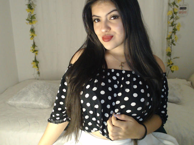 AlissonS with long black hair, wearing a black off-shoulder polka dot top, gripping her large tits, smiling with red lipstick, yellow flowers in background, white bed with pillow.
