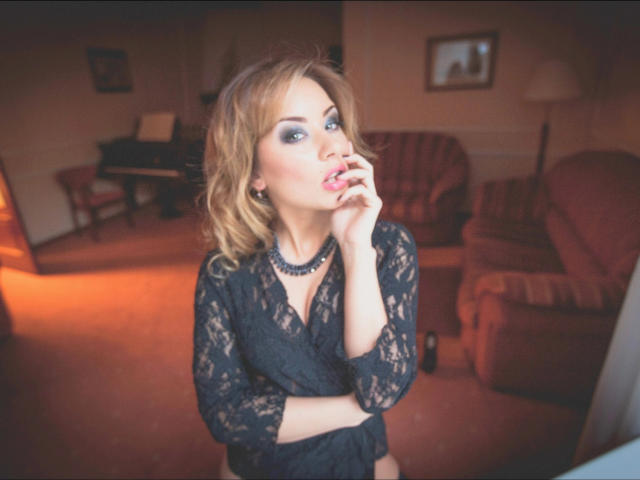 DemiMoveX with blonde hair, blue eye makeup, black lace dress, necklace, and finger in mouth, showing off her tits. Soft, warm lighting, slightly blurred background.