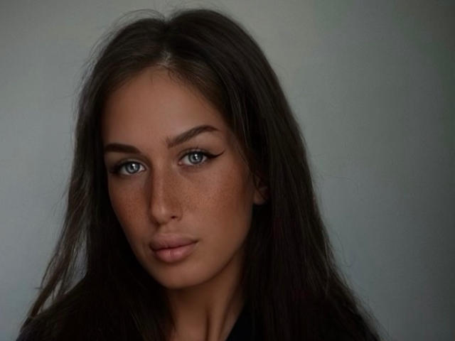 LanaXStorm heeft lang, donkerbruin haar, blauwe ogen met een vleugje eyeliner, en sproeten op haar gezicht. Ze draagt een zwarte top en heeft een serieuze uitdrukking..