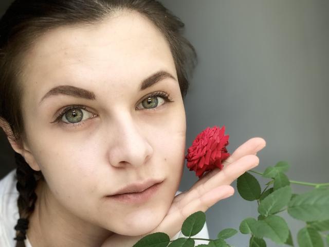 LindseyRosalyne with green eyes, fair skin, and brown hair in a braid, holding a red flower to her cheek. Her eyebrows are dark and she's wearing subtle makeup.