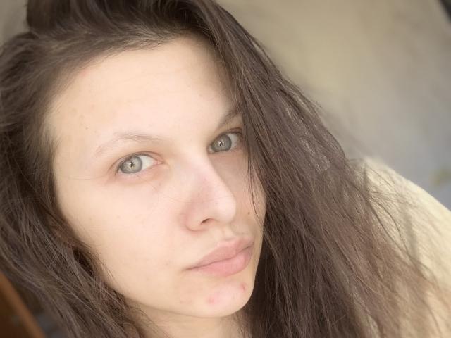 LindseyRosalyne has pale skin, green eyes, and long dark brown hair, with a small pimple on her chin, looking straight at the camera with a slight pout.