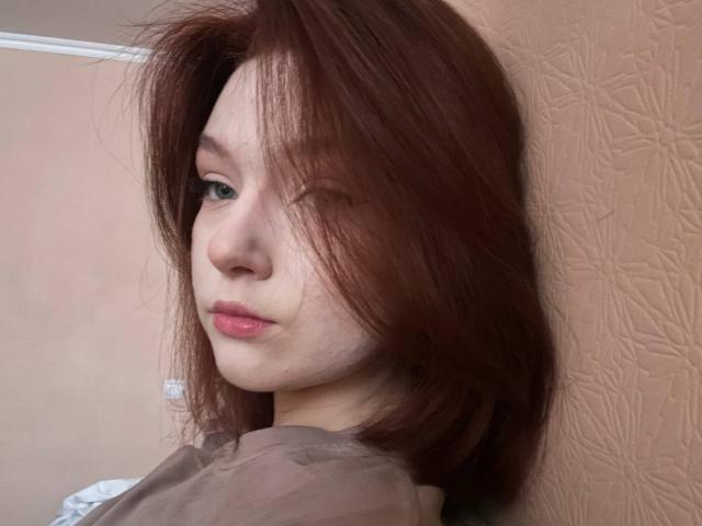 LateNightX heeft rommelig bruin haar dat één oog bedekt, bleke huid en roze lippen. Ze kijkt over haar schouder met een neutrale uitdrukking en draagt een bruin shirt..