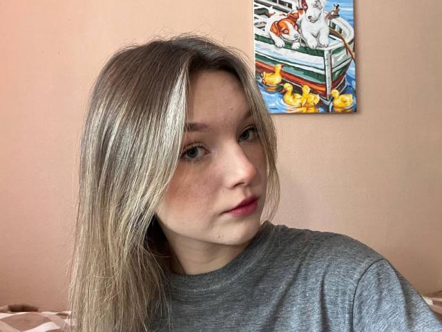LateNightX heeft blond haar, een lichte huid en blauwe ogen, draagt een grijze trui, heeft een lichte grijns, en op de achtergrond is een schilderij te zien van een witte hond met gele eendjes..