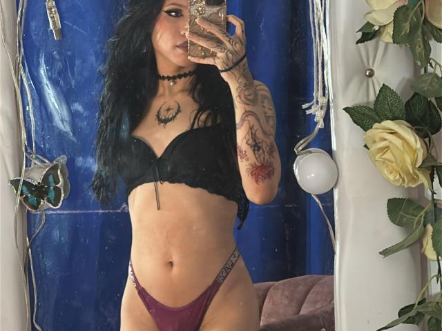 EnchantressKanne with long black hair, tattoos on arms and chest, wearing black bra and purple thong, taking mirror selfie, showing her tits and ass. Mirror reflection, blue curtain.