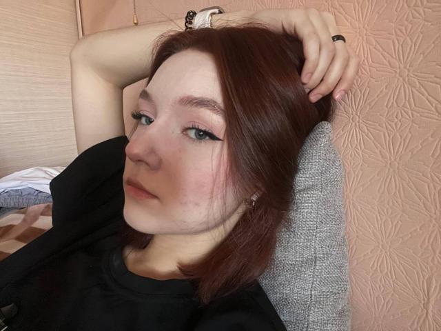 LateNightX met bleke huid, rood haar en groene ogen, gekleed in zwart, met armen omhoog, die zijwaarts kijkt met een vleugje eyeliner, een zwart shirt en een ring aan haar rechterhand..