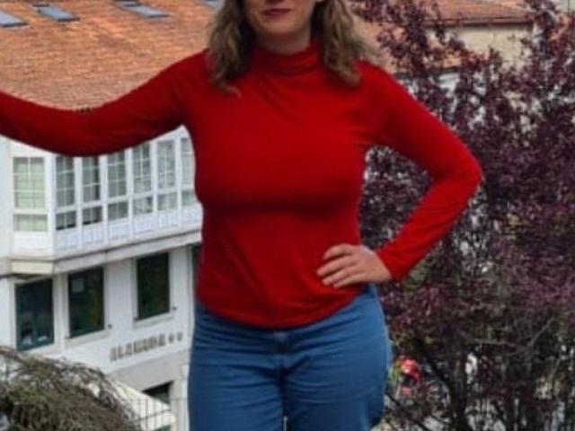 ValerieCalor-hot in a red long-sleeve top and blue jeans, standing with one arm raised, showing her tits and ass, her hand on her hip, wavy blonde hair.