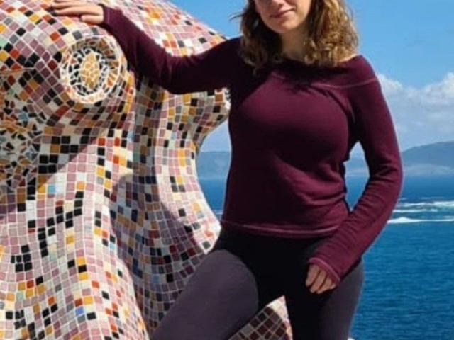 ValerieCalor-hot stands with one hand on a colorful mosaic sculpture, wearing a purple long-sleeve top and black pants, her curly brown hair, and her tits visible.