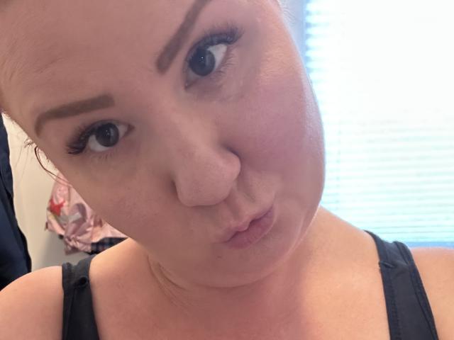 PussyDolly69 has a pouty face, slightly raised eyebrows, and thick black eyebrows. She's wearing a black tank top, showing her small tits and fair skin.