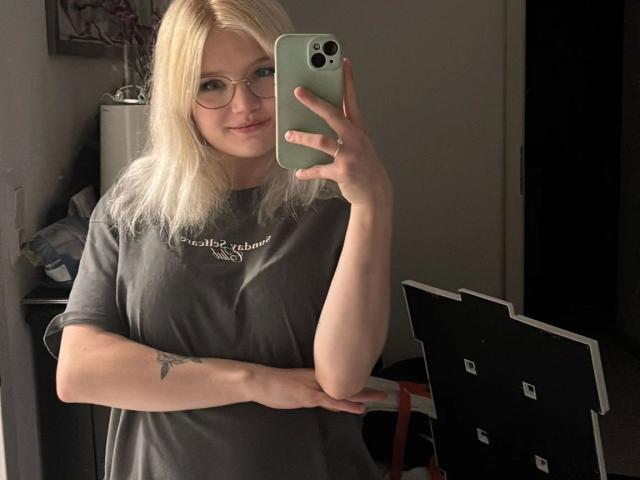 LaVani-hot with blonde hair, glasses, and a gray t-shirt, holding a phone, showing a tattoo on her arm, standing in a dimly lit room. Boobs not visible.