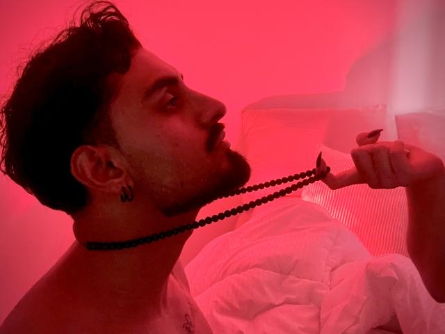 IvannaAndFriend with dark hair and beard, holding black bead necklace to mouth, red lighting, white pillow, and bed in background. Visible ear tattoo. Side profile.