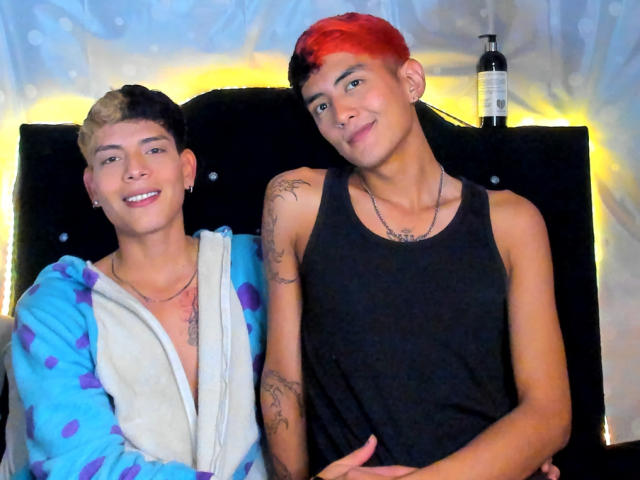 TaylorAndChris with short, dyed hair, one blonde and one red, wearing a blue hoodie and black tank top, smiling, tattoos on arm, bottle on black chair behind them.