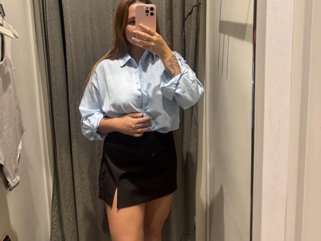 NatSorian with long brown hair, wearing a light blue shirt and black skirt, standing in a room, holding a pink phone, showing a tattoo on her right forearm.