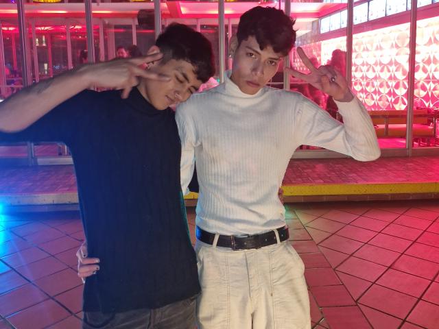 Taylor y Chris con cabello castaño corto, vistiendo un cuello alto negro y pantalones oscuros, y Taylor y Chris con cabello castaño oscuro, vistiendo un cuello alto blanco y pantalones blancos, ambos haciendo el gesto de la "V" de la paz..