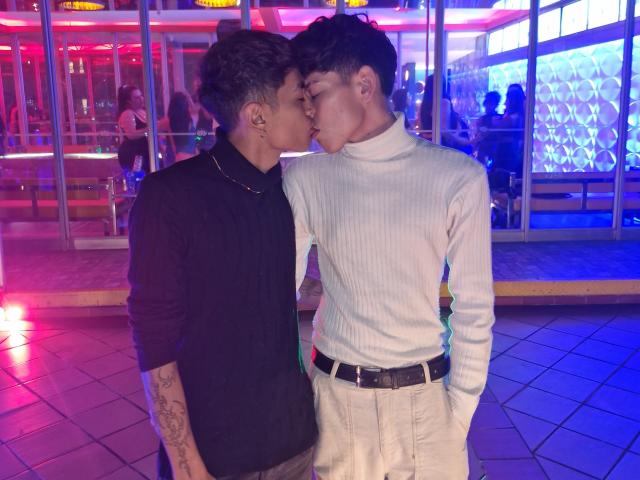 Taylor y Chris, besándose, en un club iluminado con luces de neón. Taylor tiene tatuajes, una camiseta negra y un collar. Chris lleva un suéter blanco de punto acanalado y pantalones beige. Están de pie, con los ojos cerrados..