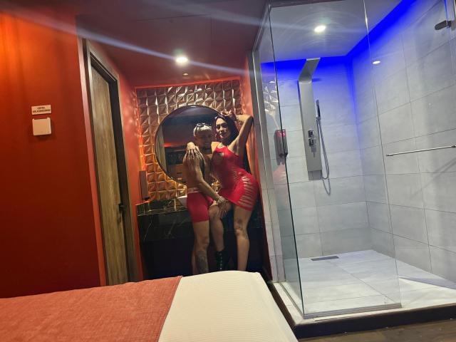 MegaSexyHot in red lingerie with a huge ass, standing in a shower with MegaSexyHot in red lingerie, grabbing their butt. Glass shower, red wall, bed in foreground.