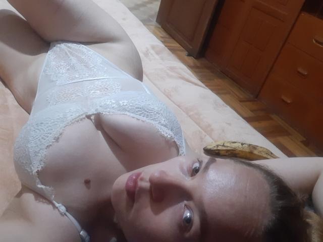 Metztona with pale skin, brown hair, and blue eyes, lying on a bed in white lace lingerie, showing her boobs and pussy. Her face is close to the camera.