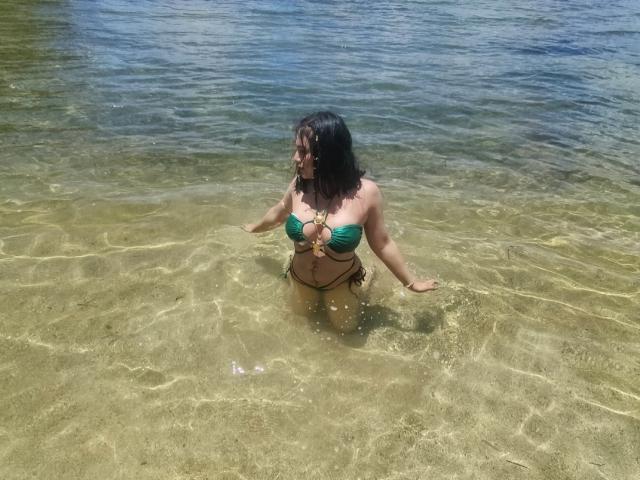 FUNKYLIE with wet dark hair, green strap bikini top, and black bikini bottoms standing in clear, shallow water, arms out, showing her tits and a small tan belly button.