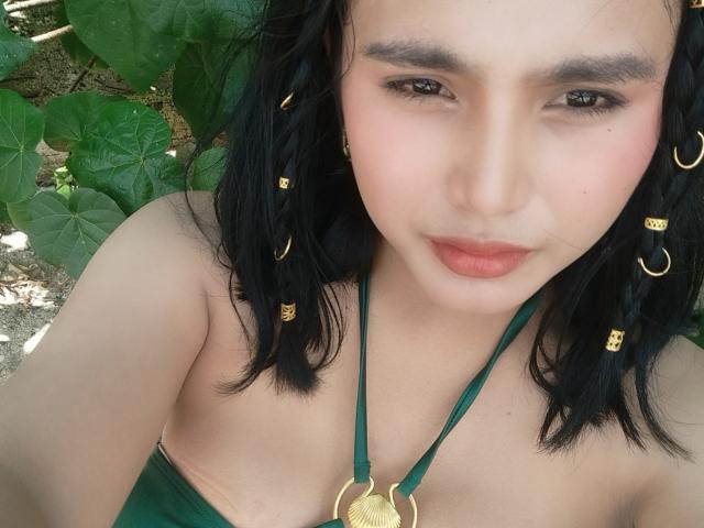 Funkylie tiene el cabello negro oscuro con cuentas doradas, ojos marrones y labios rosados. Lleva un top verde con tirantes y un colgante dorado, dejando al descubierto sus hombros..