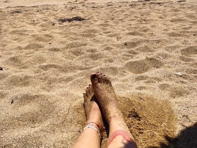 Los pies de FUNKYLIE, cubiertos de arena, con uñas de los dedos del pie marrones, cruzados a la altura de los tobillos, llevando un brazalete rosa y una pulsera blanca, en una playa soleada..