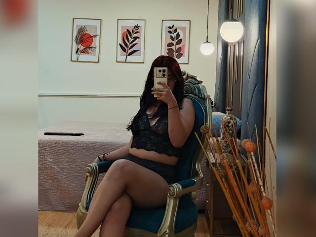 Milla Casanova, con i capelli rossi, indossa un reggiseno e una biancheria intima in pizzo nero, seduta su una sedia verde, tiene in mano un telefono, selfie allo specchio, mostrando un sedere prosperoso e piccoli seni, in una stanza accogliente..