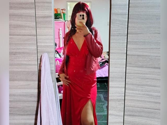 Milla Casanova, vêtue d'une robe rouge avec une fente haute, et d'une veste rouge, tenant un téléphone pour un selfie devant un miroir, montrant ses fesses et ses seins, avec des cheveux roux et un regard sexy et confiant..