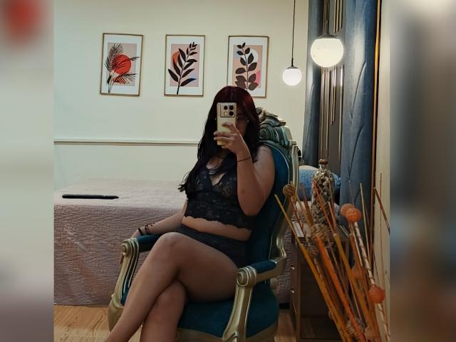 Milla Casanova, con cabello rojo, lencería de encaje negro, sentada en una silla azul, sosteniendo un teléfono, mostrando su trasero, en una habitación con arte floral y una cama..