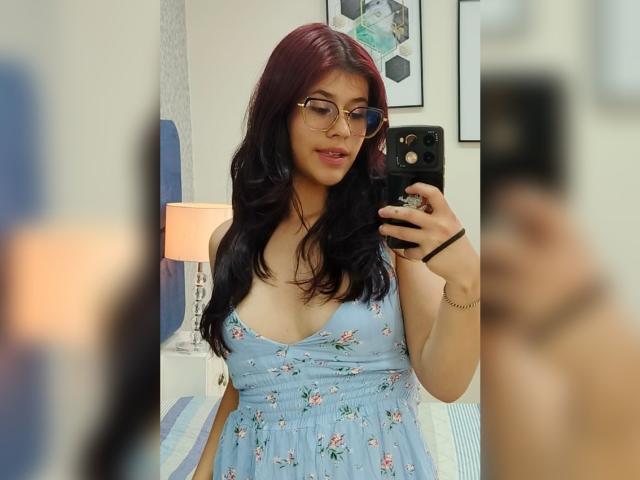 MillaCasanova with red hair, glasses, and a blue floral dress, showing her tits in a mirror selfie, holding a black phone. Her reflection is slightly blurred.