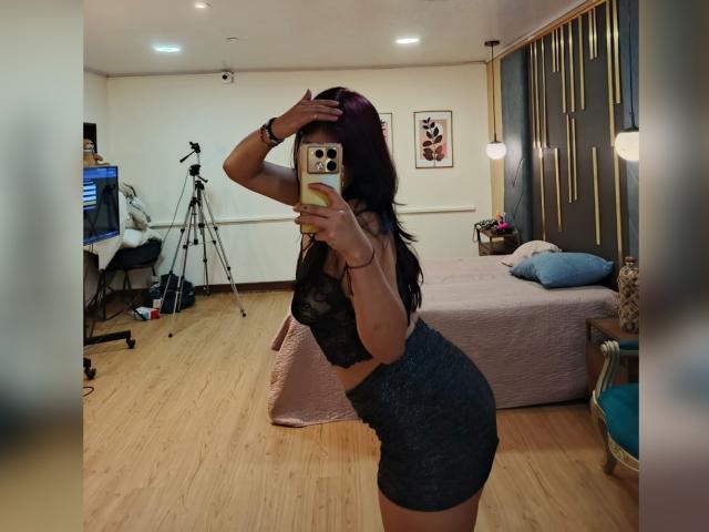 MillaCasanova with dark hair, wearing a black lace bra and gray shorts, posing sideways in a bedroom, holding a phone, showing her ass and small tits, in a mirror selfie.