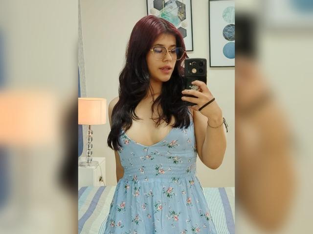 MillaCasanova with red hair, glasses, and a blue floral dress, showing her tits in a mirror selfie. She holds a black phone, standing in a bedroom.