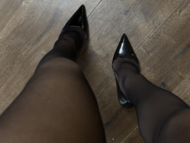 LanaXStorm's black pantyhose-covered legs in shiny black pointed-toe heels on a wooden floor, viewed from above. Boobs and pussy are not visible, just the lower half.