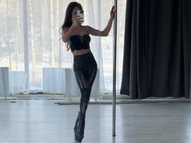 LanaXStorm, con cabello largo y castaño, un top negro de encaje sin tirantes y pantalones negros con cordones, de pie junto a un poste, tomando una selfie con un espejo, mostrando su trasero y sus pequeños pechos..