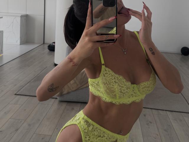 LanaXStorm trägt einen neon-gelben Spitzen-BH und Slips und zeigt ihre Brüste und Vulva in einem Spiegel-Selfie, hält ihre Nägel im Mund, hat Tattoos an den Armen, steht auf einem Holzboden und hält ein Telefon..