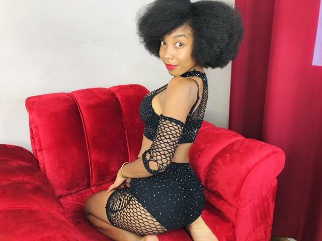 TynnaMagik with big Afro, wearing black fishnet lingerie, sitting on red velvet couch, showing her round ass and small tits, looking back with red lipstick.