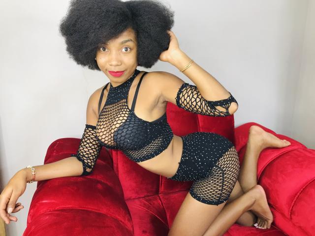 TynnaMagik with dark curly hair, wearing black fishnet lingerie with studs, on a red velvet couch, kneeling, showing her ass and small tits, with a red lipstick smile.